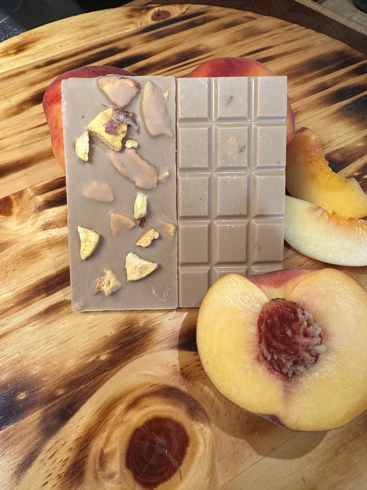 A peach studded chocolate bar on a wood background with fresh peaches in the foreground. 